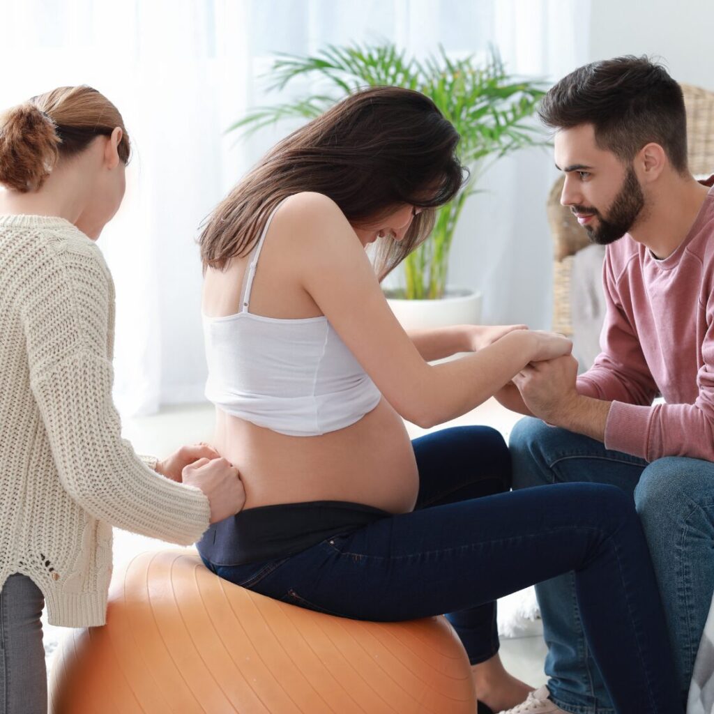 Grávida em bola de pilates sendo apoiada por acompanhante.